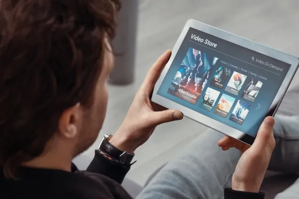 A man is seen from over the shoulder holding a tablet displaying a "Video Store" screen with movie thumbnails and genre categories, labeled "Video On Demand."